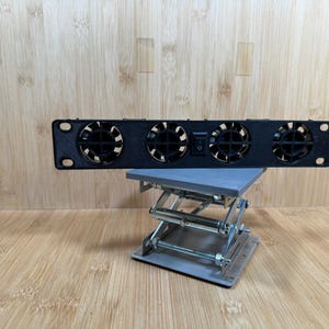 May include: A black cooling fan with four circular fans and an on/off switch. The fan is mounted on a gray scissor lift. The background is a light brown wooden surface.