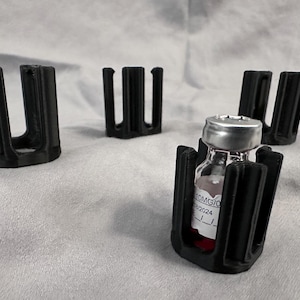 May include: Four black 3D printed vial holders, three empty and one holding a clear glass vial with a silver cap and a red liquid. The vial label reads "10MG/1mL EXP 2024".