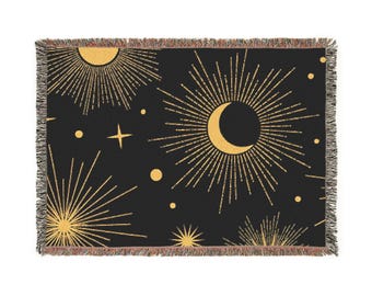 May include: Black throw blanket with a celestial design featuring gold suns, moons, stars, and dots. The blanket has a fringed edge. The celestial pattern is a repeating design across the blanket.