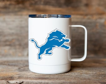 May include: White travel mug with a blue Detroit Lions logo featuring a lion. The mug has a lid and a handle.