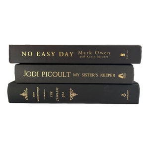 May include: A stack of three black hardcover books with gold lettering. The top book is titled "No Easy Day" by Mark Owen with Kevin Maurer. The middle book is titled "My Sister's Keeper" by Jodi Picoult. The bottom book is titled "The Starless Sea" by Erin Morgenstern.