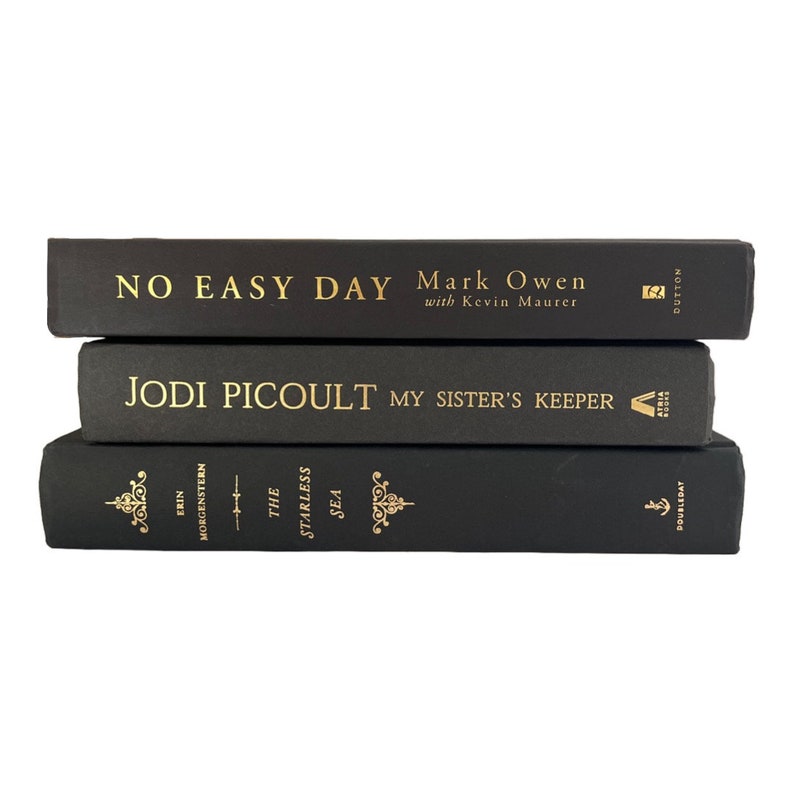 May include: A stack of three black hardcover books with gold lettering. The top book is titled "No Easy Day" by Mark Owen with Kevin Maurer. The middle book is titled "My Sister's Keeper" by Jodi Picoult. The bottom book is titled "The Starless Sea" by Erin Morgenstern.