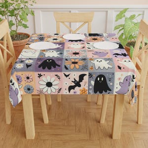 May include: A square tablecloth featuring a repeating grid pattern of Halloween-themed illustrations. The design includes white, black, and purple ghosts, black bats, orange pumpkins, and various colourful daisy flowers on pastel backgrounds.