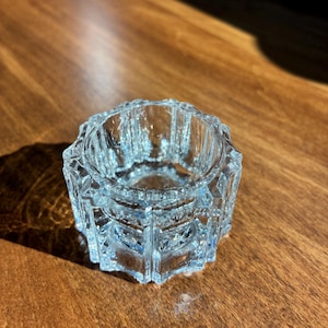 May include: A clear, faceted glass votive candle holder. The holder has a star-shaped base and a circular opening. The glass has a slight blue tint, and the facets catch the light, creating a sparkling effect. The candle holder is likely vintage.