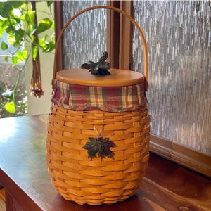 May include: A round woven basket with a light brown finish, featuring a wooden lid topped with a dark metal leaf handle. The basket has a plaid fabric liner in red, green, and tan, and a single arched handle. A dark metal leaf accent is attached.