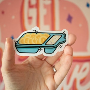 May include: A blue and white sticker of a divided container with yellow noodles in one section and white frosting with sprinkles in the other section. The sticker has a sparkly design.