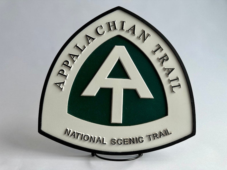 K&ouml;nnte beinhalten: Ein wei&szlig;es und schwarzes Schild mit gr&uuml;nem Hintergrund. Das Schild ist dreieckig mit abgerundeten Ecken. Auf dem Schild steht "Appalachian Trail National Scenic Trail" und es hat ein wei&szlig;es "A" mit einem wei&szlig;en "T" darin.
