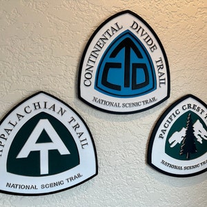 K&ouml;nnte beinhalten: Drei dreieckige Schilder mit schwarzen R&auml;ndern und wei&szlig;en Hintergr&uuml;nden. Jedes Schild zeigt das Logo eines anderen National Scenic Trail: den Continental Divide Trail, den Appalachian Trail und den Pacific Crest Trail. Die Logos sind in Gr&uuml;n, Blau und Schwarz.