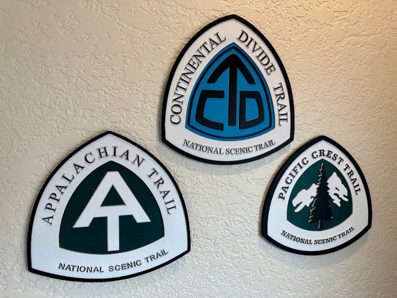 K&ouml;nnte beinhalten: Drei dreieckige Schilder mit schwarzen R&auml;ndern und wei&szlig;en Hintergr&uuml;nden. Jedes Schild zeigt das Logo eines anderen National Scenic Trail: den Continental Divide Trail, den Appalachian Trail und den Pacific Crest Trail. Die Logos sind in Gr&uuml;n, Blau und Schwarz.