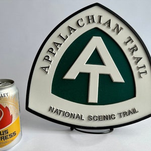 K&ouml;nnte beinhalten: Ein wei&szlig;es und gr&uuml;nes Schild mit schwarzer Beschriftung, auf dem "Appalachian Trail National Scenic Trail" steht. Das Schild zeigt ein wei&szlig;es "A" mit einem wei&szlig;en Querbalken auf gr&uuml;nem Hintergrund. Das Schild steht auf einem schwarzen St&auml;nder.