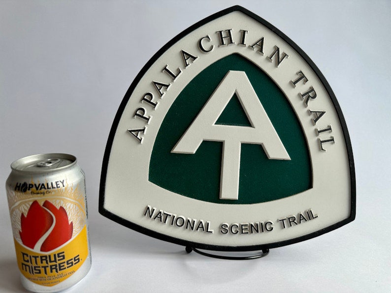 K&ouml;nnte beinhalten: Ein wei&szlig;es und gr&uuml;nes Schild mit schwarzer Beschriftung, auf dem "Appalachian Trail National Scenic Trail" steht. Das Schild zeigt ein wei&szlig;es "A" mit einem wei&szlig;en Querbalken auf gr&uuml;nem Hintergrund. Das Schild steht auf einem schwarzen St&auml;nder.
