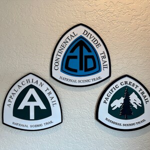 K&ouml;nnte beinhalten: Drei wei&szlig;e, schwarze und gr&uuml;ne dreieckige Schilder mit den Logos des Continental Divide Trail, Appalachian Trail und Pacific Crest Trail. Jedes Schild tr&auml;gt die Aufschrift "National Scenic Trail".
