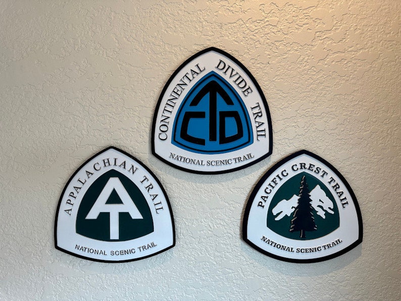 K&ouml;nnte beinhalten: Drei wei&szlig;e, schwarze und gr&uuml;ne dreieckige Schilder mit den Logos des Continental Divide Trail, Appalachian Trail und Pacific Crest Trail. Jedes Schild tr&auml;gt die Aufschrift "National Scenic Trail".