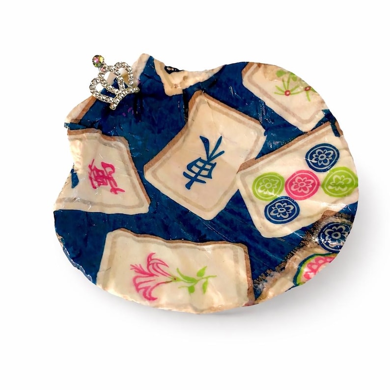 May include: A decorative, round object with a dark blue background and several off-white tiles. The tiles feature colorful designs and Chinese characters. A small, jeweled crown is attached to the top. The object has a unique, handmade appearance.