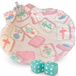 May include: A decorative seashell-shaped tray with floral and bird illustrations on playing card-like designs. A small jeweled crown sits on the edge. Two turquoise dice with white dots are in the foreground.