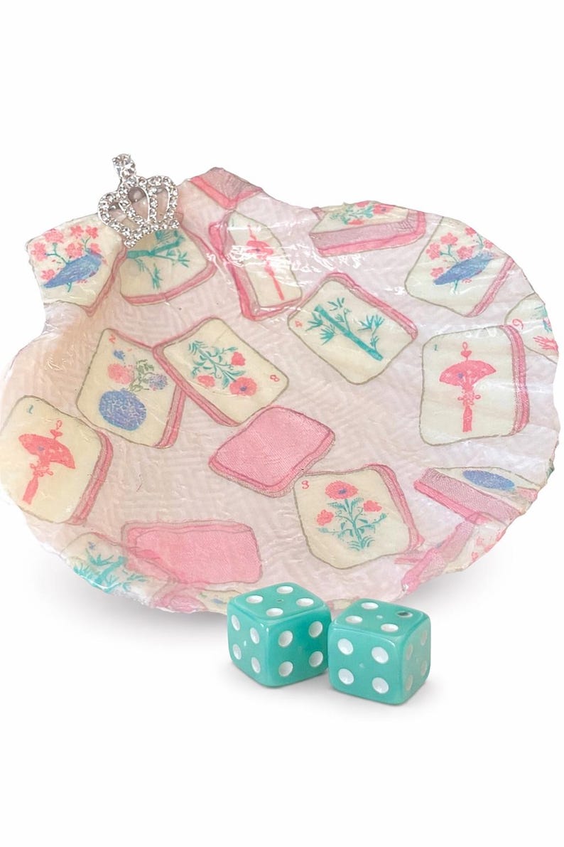 May include: A decorative seashell-shaped tray with floral and bird illustrations on playing card-like designs. A small jeweled crown sits on the edge. Two turquoise dice with white dots are in the foreground.