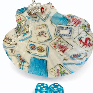 May include: A decorative seashell-shaped dish with a playing card design, a small jeweled crown, and two blue dice. The dish features a white background with blue and red accents, and the dice are transparent.