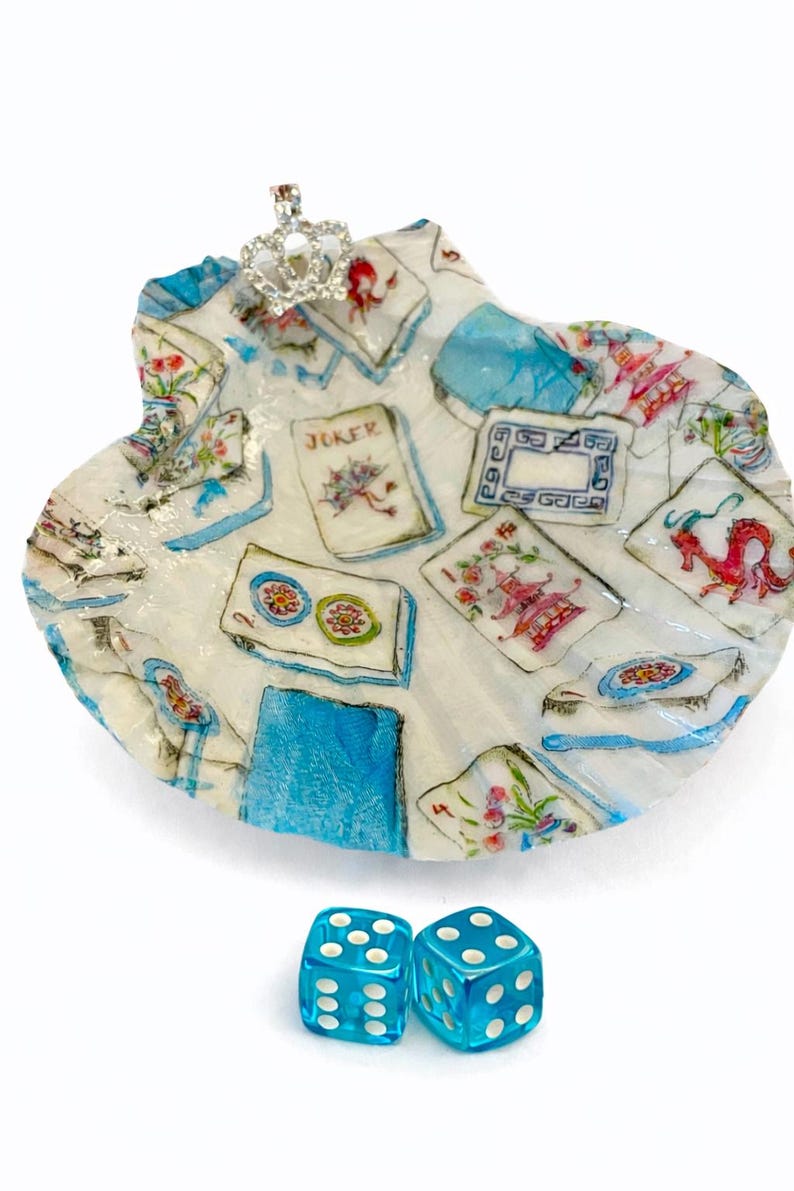 May include: A decorative seashell-shaped dish with a playing card design, a small jeweled crown, and two blue dice. The dish features a white background with blue and red accents, and the dice are transparent.