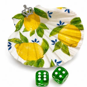 May include: A white seashell painted with yellow lemons, green leaves, and blue accents. A small silver crown is attached to the shell. Two green dice with white dots are below the shell.