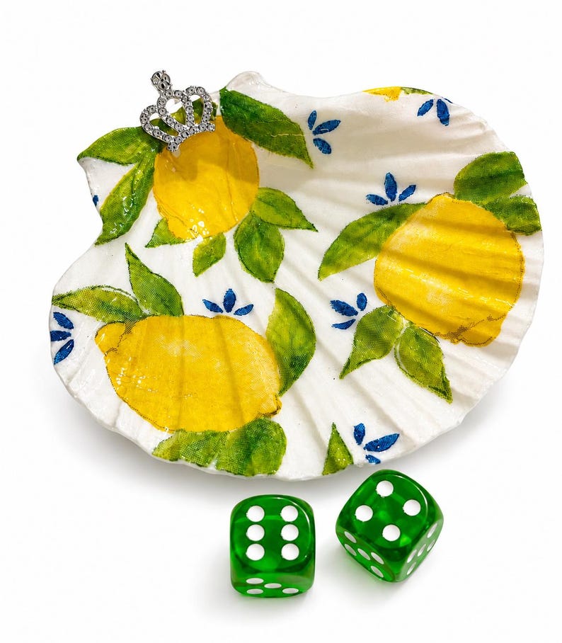 May include: A white seashell painted with yellow lemons, green leaves, and blue accents. A small silver crown is attached to the shell. Two green dice with white dots are below the shell.