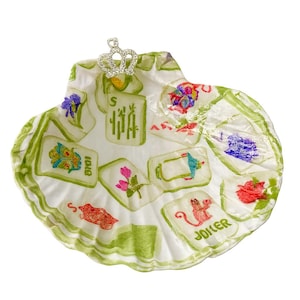 May include: A decorative seashell-shaped dish with a white base and green trim. The dish features colorful illustrations of playing cards and a small silver crown. The word "JOKER" is visible on one of the cards.
