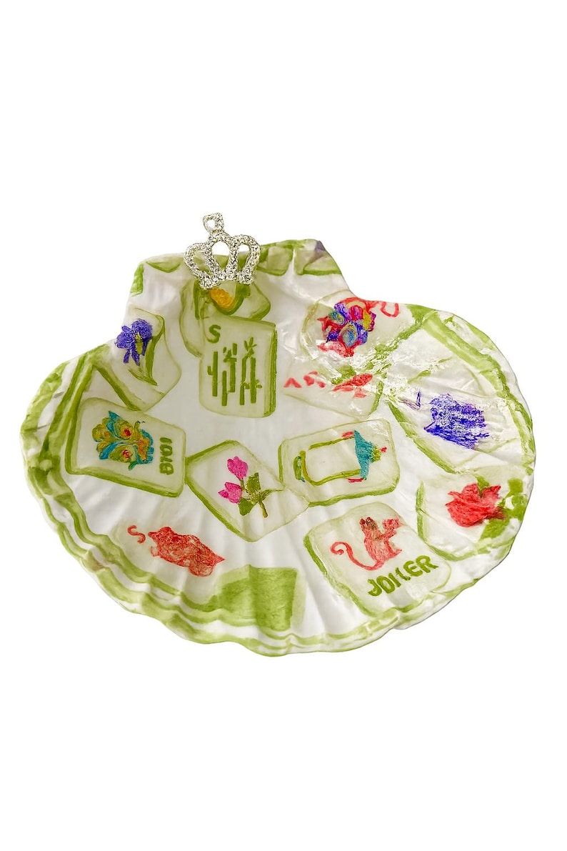May include: A decorative seashell-shaped dish with a white base and green trim. The dish features colorful illustrations of playing cards and a small silver crown. The word "JOKER" is visible on one of the cards.