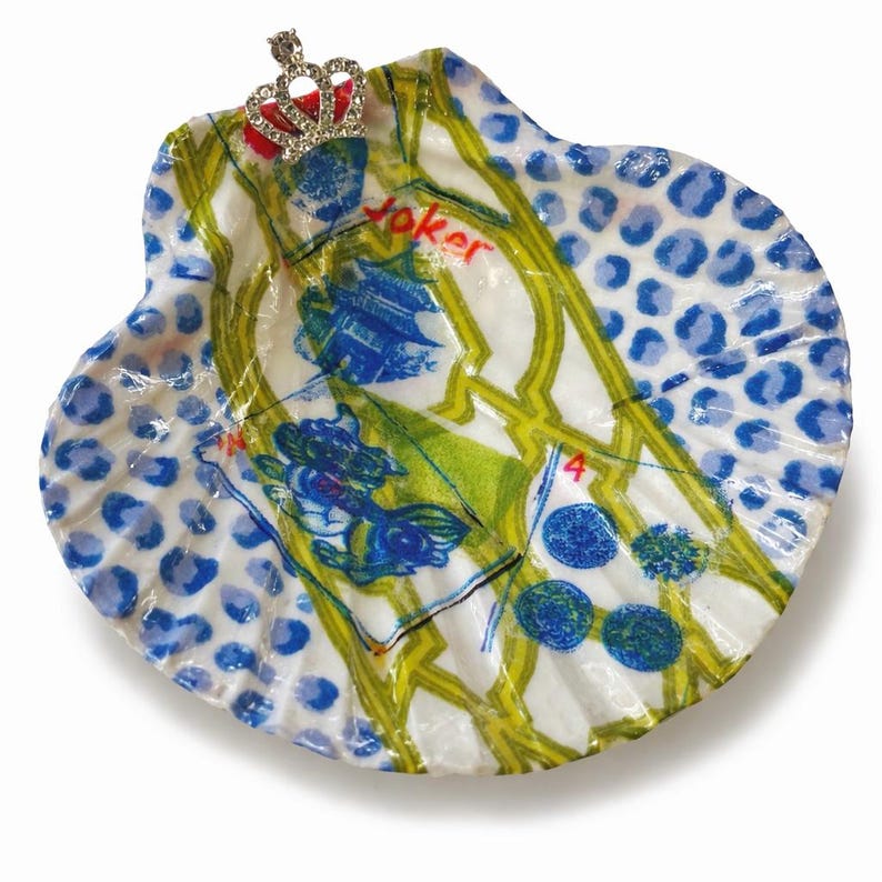 May include: A decorative seashell with a playing card design, featuring blue, green, and yellow accents. The shell is topped with a small, jeweled crown. The word "Joker" is visible on the shell.
