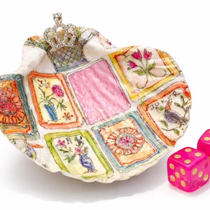 May include: A seashell-shaped decorative dish with a crown on top, featuring a painted playing card design. Two pink dice with yellow dots are next to the dish. The dish has a pink center and colorful floral and bird illustrations.