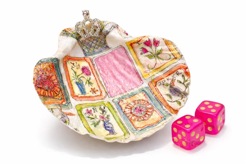 May include: A seashell-shaped decorative dish with a crown on top, featuring a painted playing card design. Two pink dice with yellow dots are next to the dish. The dish has a pink center and colorful floral and bird illustrations.