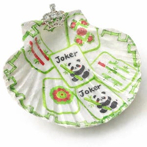 May include: A white seashell decorated with a hand-painted Mahjong game design. The design includes green bamboo, red flowers, and panda bear "Joker" tiles. A small, jeweled crown is attached to the top of the shell.