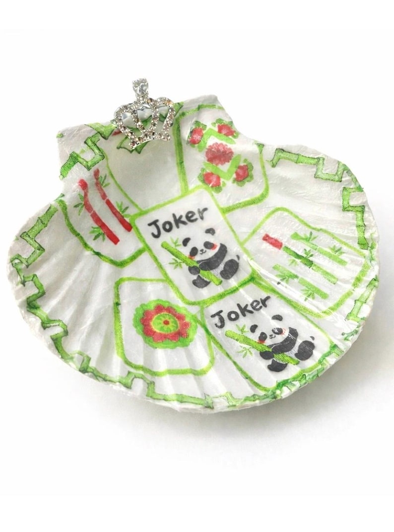 May include: A white seashell decorated with a hand-painted Mahjong game design. The design includes green bamboo, red flowers, and panda bear "Joker" tiles. A small, jeweled crown is attached to the top of the shell.