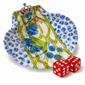 May include: A decorative seashell-shaped dish with a playing card design, featuring the word "Joker" and a small jeweled crown. Two red dice with white dots are placed next to the dish. The dish has blue and white polka dots.