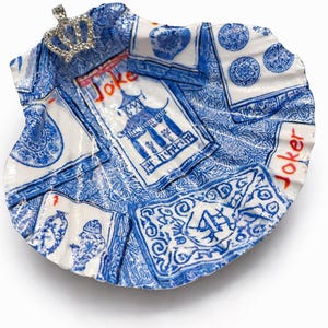 May include: A decorative, shell-shaped dish with a blue and white playing card design. The dish features a small, jeweled crown and the word "Joker" in red. The design includes images of pagodas and card suits.