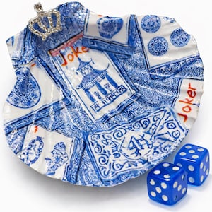 May include: A decorative, shell-shaped dish with a blue and white playing card design, featuring the word "Joker" and a small jeweled crown. Two blue dice with white dots are next to the dish.