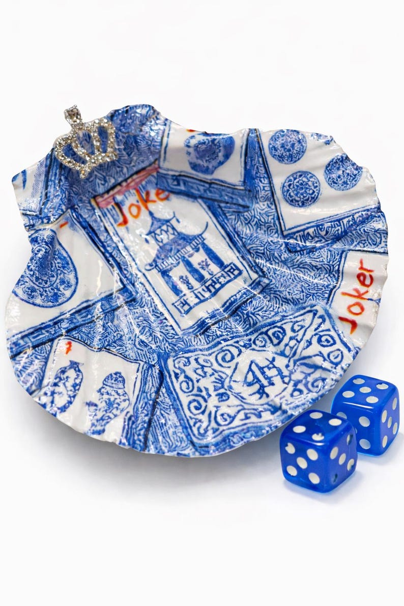 May include: A decorative, shell-shaped dish with a blue and white playing card design, featuring the word "Joker" and a small jeweled crown. Two blue dice with white dots are next to the dish.