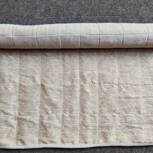 K&ouml;nnte beinhalten: Ein zusammengerollter wei&szlig;er Baumwoll-Teppich mit einem dezenten Fischgr&auml;tenmuster. Der Teppich liegt auf einem grauen Teppich.
