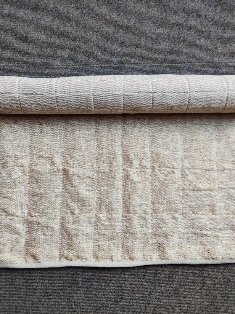 K&ouml;nnte beinhalten: Ein zusammengerollter wei&szlig;er Baumwoll-Teppich mit einem dezenten Fischgr&auml;tenmuster. Der Teppich liegt auf einem grauen Teppich.