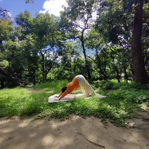 K&ouml;nnte beinhalten: Eine Person, die Yoga in einer Downward-Facing-Dog-Pose auf einer wei&szlig;en Yogamatte in einer Waldumgebung macht. Die Person tr&auml;gt ein orangefarbenes Shirt und eine wei&szlig;e Hose. Der Wald ist &uuml;ppig und gr&uuml;n mit hohen B&auml;umen und einem blauen Himmel mit wei&szlig;en Wolken.
