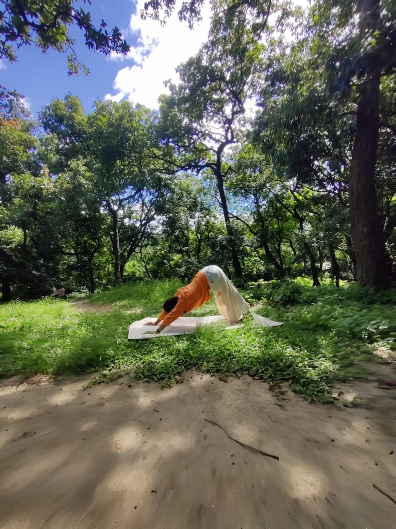 K&ouml;nnte beinhalten: Eine Person, die Yoga in einer Downward-Facing-Dog-Pose auf einer wei&szlig;en Yogamatte in einer Waldumgebung macht. Die Person tr&auml;gt ein orangefarbenes Shirt und eine wei&szlig;e Hose. Der Wald ist &uuml;ppig und gr&uuml;n mit hohen B&auml;umen und einem blauen Himmel mit wei&szlig;en Wolken.