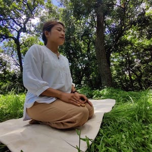 K&ouml;nnte beinhalten: Eine Person sitzt in meditativer Haltung auf einer wei&szlig;en Yogamatte mit dem Wort "Zen" darauf gedruckt. Die Matte liegt auf einem Bett aus gr&uuml;nem Gras. Die Person tr&auml;gt ein wei&szlig;es Hemd und eine braune Hose. Der Hintergrund ist ein &uuml;ppiger gr&uuml;ner Wald mit hohen B&auml;umen.