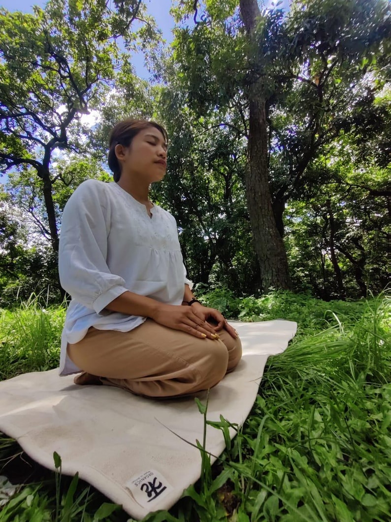 K&ouml;nnte beinhalten: Eine Person sitzt in meditativer Haltung auf einer wei&szlig;en Yogamatte mit dem Wort "Zen" darauf gedruckt. Die Matte liegt auf einem Bett aus gr&uuml;nem Gras. Die Person tr&auml;gt ein wei&szlig;es Hemd und eine braune Hose. Der Hintergrund ist ein &uuml;ppiger gr&uuml;ner Wald mit hohen B&auml;umen.