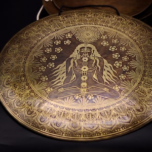 May include: A large, round, brass gong with a gold mandala design. The centre of the gong features a stylised image of a person in a seated meditation pose with seven chakras depicted on their torso.