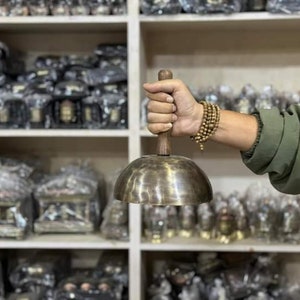 May include: A person holds a large, round, brass singing bowl with a wooden handle. The bowl is in focus, and the person's hand and arm are blurred. The bowl is surrounded by shelves of similar bowls and other items.