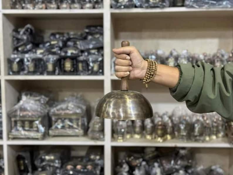 May include: A person holds a large, round, brass singing bowl with a wooden handle. The bowl is in focus, and the person's hand and arm are blurred. The bowl is surrounded by shelves of similar bowls and other items.