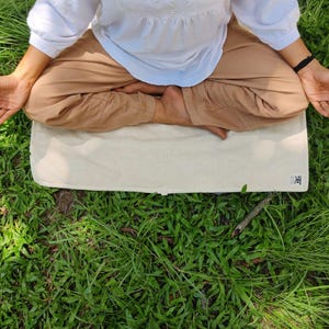 K&ouml;nnte beinhalten: Eine Person sitzt in meditativer Haltung auf einer wei&szlig;en Yogamatte mit dem Wort "禅" darauf. Die Matte liegt auf einem Bett aus gr&uuml;nem Gras.