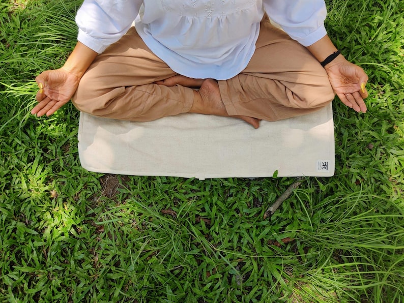 K&ouml;nnte beinhalten: Eine Person sitzt in meditativer Haltung auf einer wei&szlig;en Yogamatte mit dem Wort "禅" darauf. Die Matte liegt auf einem Bett aus gr&uuml;nem Gras.