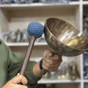 May include: A person is holding a large, antique-looking, brass singing bowl with a wooden mallet with a blue felt head.