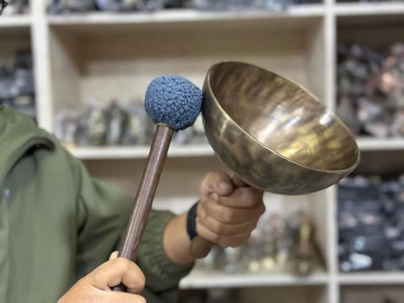 May include: A person holds a large, antique-looking, brass singing bowl with a wooden mallet with a blue felt head.