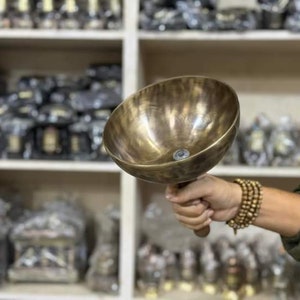 May include: A large, round, brass singing bowl with a wooden handle. The bowl has a hammered texture and a dark brown patina.