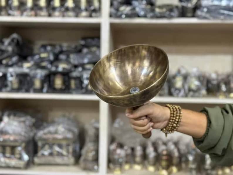 May include: A large, round, brass singing bowl with a wooden handle. The bowl has a hammered texture and a dark brown patina.